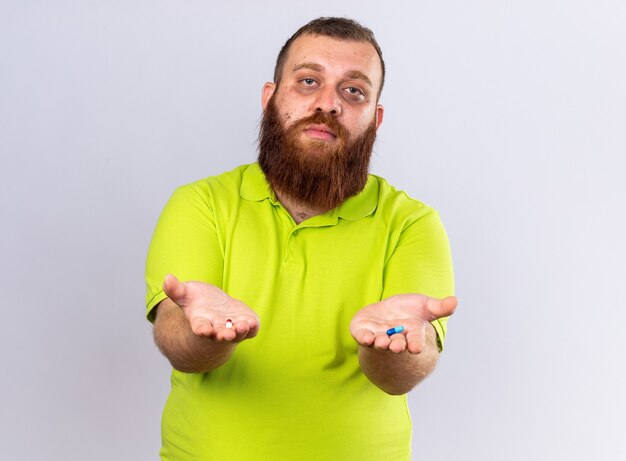 Homme barbu malsain en polo jaune tenant différentes pilules se sentant malade souffrant de la grippe debout sur un mur blanc