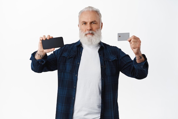 Homme barbu mature sérieux montrant l'écran du téléphone portable et la carte de crédit montrant quelque chose sur l'écran du smartphone debout sur fond blanc Copier l'espace