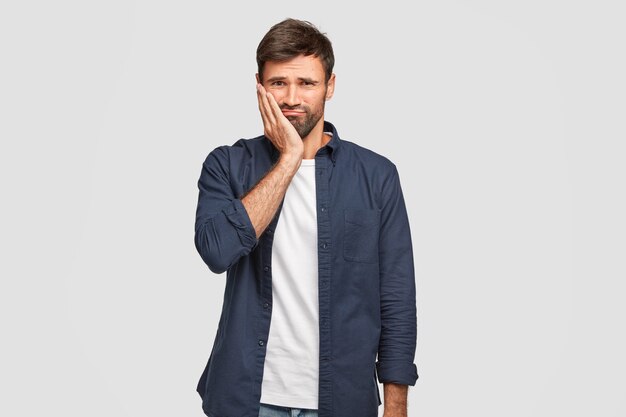 Un homme barbu mécontent garde la main sur la joue, fronce les sourcils avec mécontentement, entend des nouvelles négatives, ne veut pas aller au travail, porte une chemise bleu foncé à la mode, se tient seul contre un mur blanc