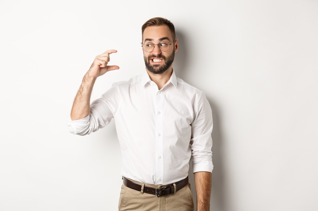 Homme barbu mécontent montrant un petit geste, l'air déçu, debout contre le blanc