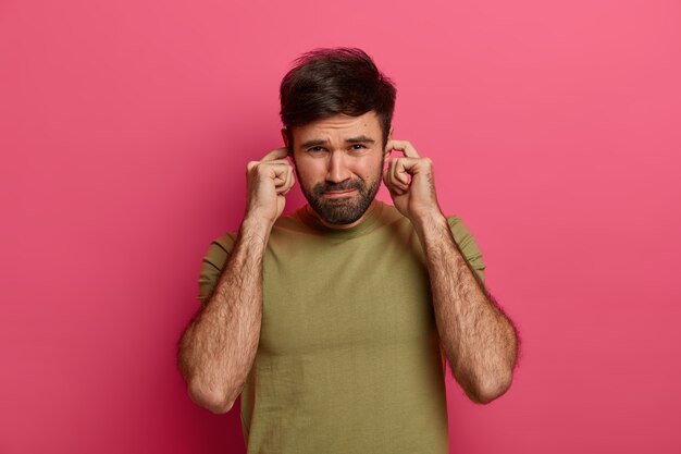 Un homme barbu mécontent ne veut pas écouter la conversation entre amis, bouche les trous d'oreille avec les doigts, ne peut pas se concentrer dans une atmosphère bruyante, évite les sons désagréables, habillé en t-shirt décontracté, pose à l'intérieur