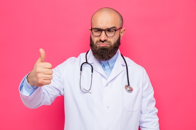 Photo gratuite homme barbu médecin en blouse blanche avec stéthoscope autour du cou portant des lunettes à côté avec un visage sérieux montrant les pouces vers le haut