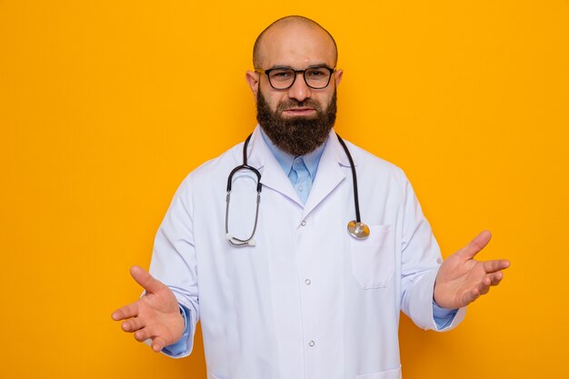 Homme barbu médecin en blouse blanche avec stéthoscope autour du cou portant des lunettes regardant la caméra souriant confiant heureux et positif levant les bras debout sur fond orange