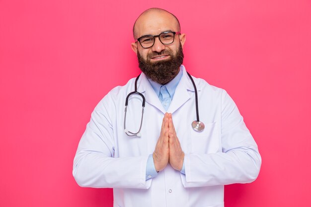 Homme barbu médecin en blouse blanche avec stéthoscope autour du cou portant des lunettes à sourire joyeusement tenant les mains ensemble comme le geste de namaste