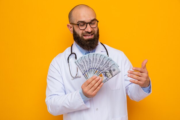 Homme barbu médecin en blouse blanche avec stéthoscope autour du cou portant des lunettes tenant de l'argent heureux et heureux souriant