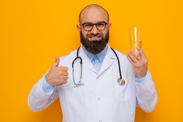 Homme barbu médecin en blouse blanche avec stéthoscope autour du cou portant des lunettes tenant un verre d'eau regardant la caméra souriant joyeusement montrant les pouces vers le haut debout sur fond orange