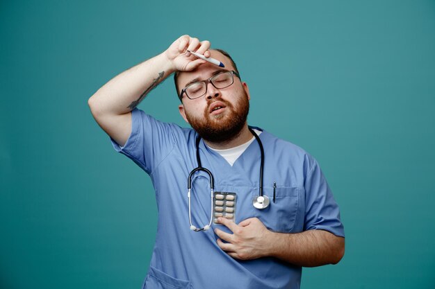 Homme barbu médecin en uniforme avec stéthoscope autour du cou portant des lunettes tenant un thermomètre et des pilules à l'air fatigué et surmené debout sur fond bleu