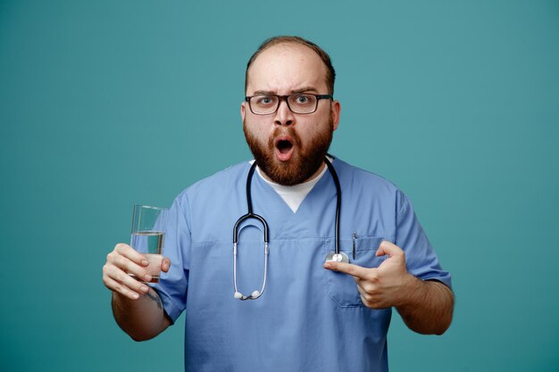 Homme barbu médecin en uniforme avec stéthoscope autour du cou portant des lunettes tenant un verre d'eau regardant la caméra étonné et surpris debout sur fond bleu