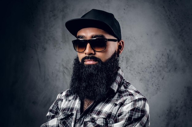 Homme barbu noir pensif vêtu d'une chemise polaire, de lunettes de soleil et d'une casquette de baseball posant sur fond gris.