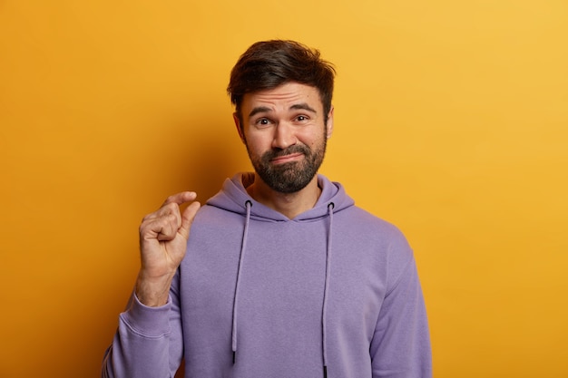 Un homme barbu non impressionné montre un petit geste, montre une petite quantité de quelque chose, façonne un petit objet, dit que c'est tout ce dont j'ai besoin, vêtu d'un sweat-shirt décontracté, isolé sur un mur jaune