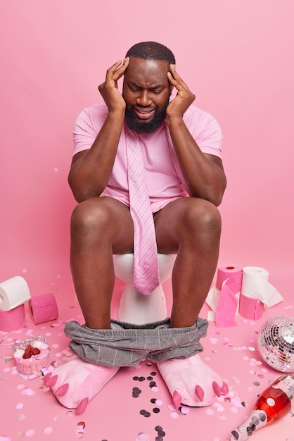 Photo gratuite l'homme barbu à la peau foncée souffre de maux de tête et se sent mal a la gueule de bois après la pose de la fête sur la cuvette des toilettes dans les toilettes entouré d'objets laissés après la célébration isolés sur fond rose.