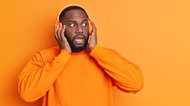 Photo gratuite un homme barbu à la peau sombre regarde les yeux obstrués garde les mains sur des écouteurs stéréo écoute la musique réagit à quelque chose