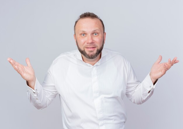 Homme barbu portant une chemise blanche confus répandant les bras sur le côté n'ayant pas de réponse debout sur un mur blanc