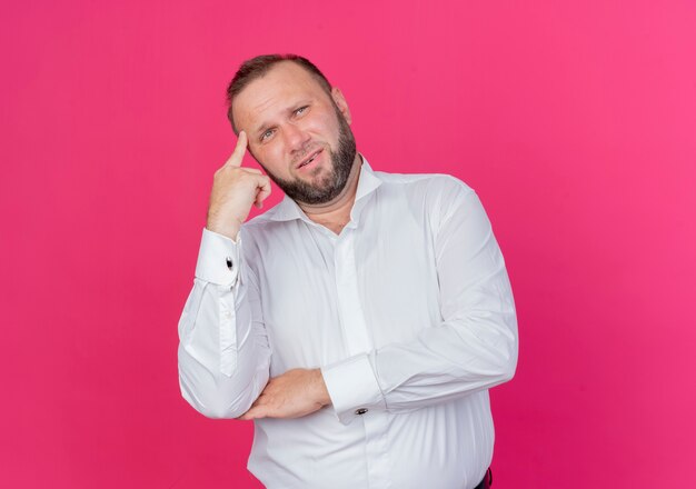 Homme barbu portant une chemise blanche à côté perplexe debout sur un mur rose