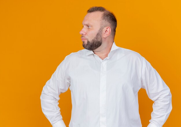 Homme barbu portant une chemise blanche à côté avec un visage sérieux debout sur un mur orange