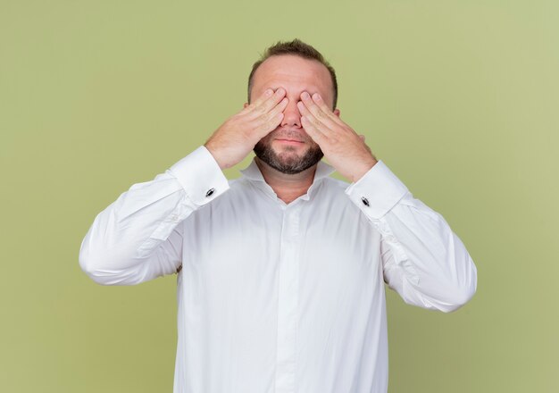 Homme barbu portant une chemise blanche couvrant les yeux avec les bras debout sur le mur léger