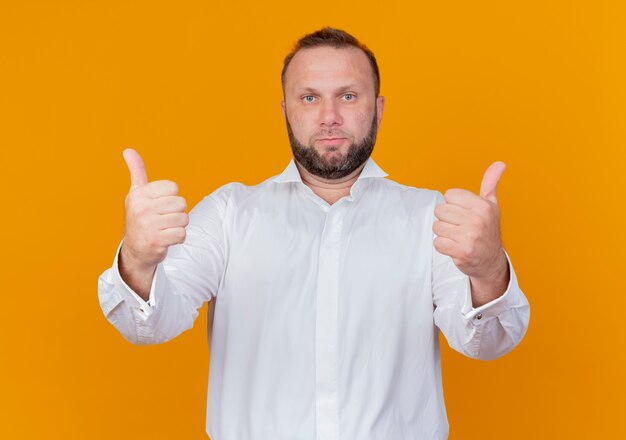 Homme barbu portant une chemise blanche avec une expression confiante montrant les pouces vers le haut debout sur un mur orange