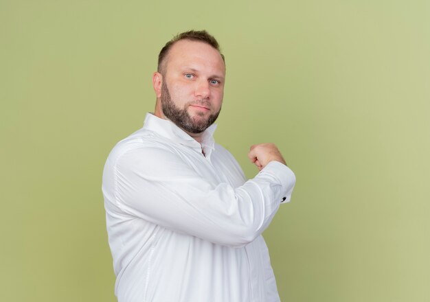 Homme barbu portant une chemise blanche avec une expression confiante pointant vers l'arrière debout sur un mur léger