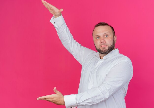 Homme barbu portant une chemise blanche montrant un geste de grande taille avec des mains mesure symbole debout sur un mur rose