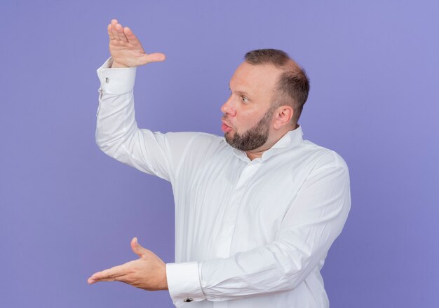 Homme barbu portant une chemise blanche montrant un geste de grande taille avec les mains à la mesure de symbole surpris et étonné debout sur le mur bleu