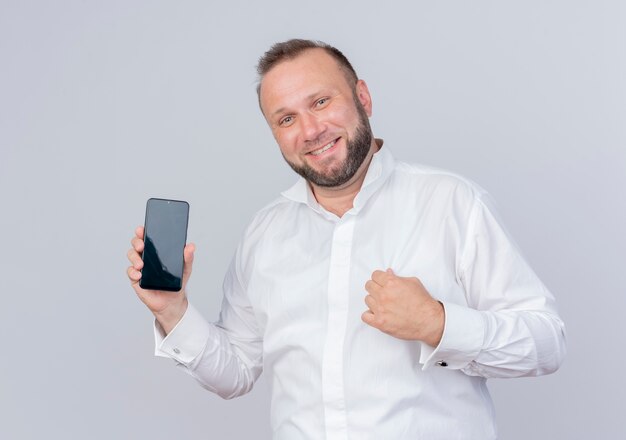 Homme barbu portant une chemise blanche montrant smartphone poing serrant heureux et excité debout sur un mur blanc