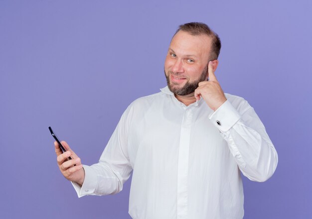 Homme barbu portant une chemise blanche parlant au téléphone mobile à la recherche de temple de pointage souriant concentré sur une tâche debout sur le mur bleu