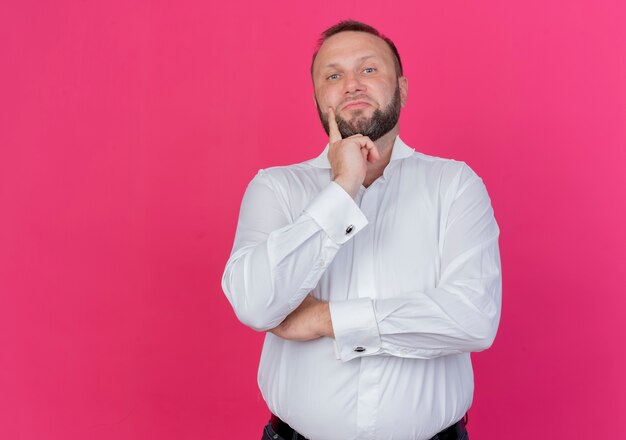 Homme barbu portant une chemise blanche à la perplexité debout sur un mur rose
