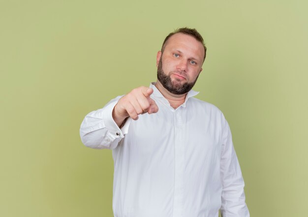Homme barbu portant une chemise blanche pointant avec l'index debout sur un mur léger