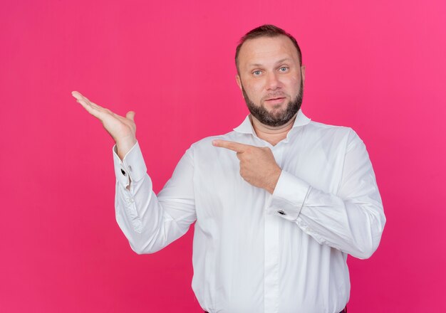 Homme barbu portant une chemise blanche présentant quelque chose avec le bras pointant avec le doigt sur le côté debout sur un mur rose