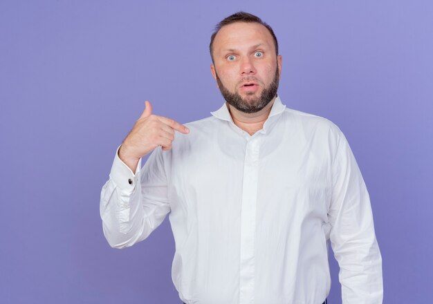 Homme barbu portant une chemise blanche à la recherche de se montrer confus et surpris debout sur un mur bleu