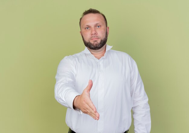 Homme barbu portant une chemise blanche salutation offrant la main debout sur un mur léger