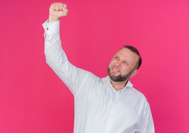 Homme barbu portant une chemise blanche serrant le poing heureux et excité se réjouissant de son succès debout sur le mur rose