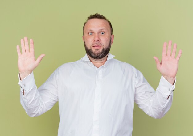 Homme barbu portant une chemise blanche soulevant des paumes dans la reddition looing confus debout sur mur léger