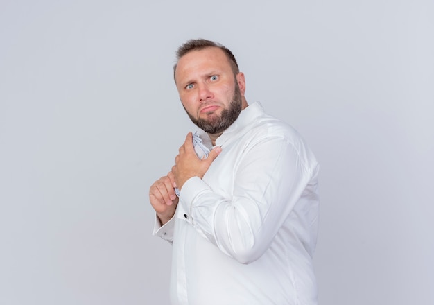 Homme barbu portant une chemise blanche tenant de l'argent cachant de l'argent avec une expression sceptique debout sur un mur blanc