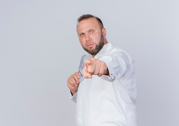 Homme barbu portant une chemise blanche tenant de l'argent cachant de l'argent pointign avec l'index pour vous debout sur un mur blanc