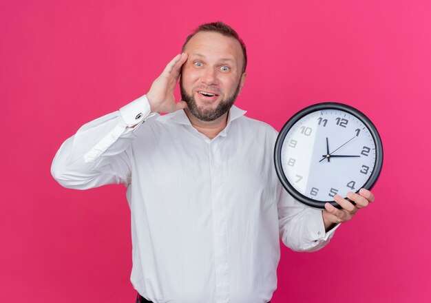 Homme barbu portant une chemise blanche tenant une horloge murale à la recherche de sourire confus debout sur un mur rose