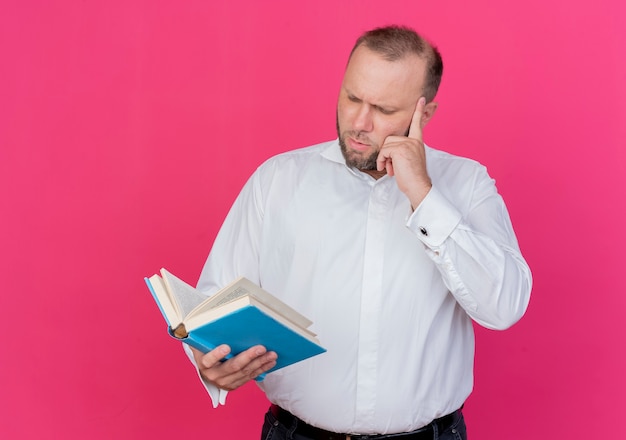 Homme barbu portant une chemise blanche tenant un livre ouvert en le regardant avec un visage sérieux debout sur un mur rose