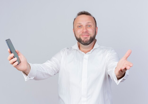 Homme barbu portant une chemise blanche tenant un smartphone faisant un geste de bienvenue avec la main souriant debout sur un mur blanc