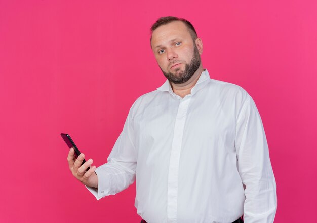 Homme barbu portant une chemise blanche tenant un smartphone avec un visage sérieux debout sur un mur rose