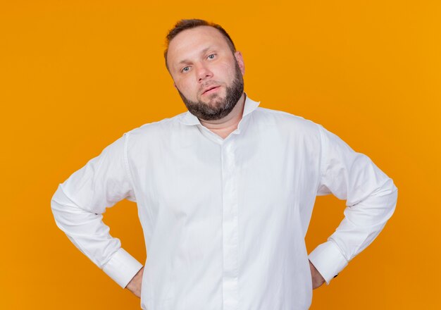 Homme barbu portant une chemise blanche avec un visage sérieux avec les bras à la hanche debout sur le mur orange