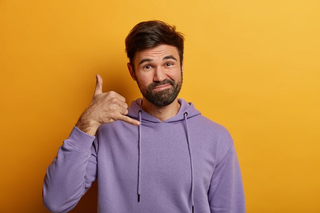 Un homme barbu à la recherche amicale et gai fait un geste de rappel, fait semblant de tenir le téléphone près de l'oreille, imite l'appel et la communication mobile, montre un signe de numérotation, porte un sweat à capuche, pose à l'intérieur