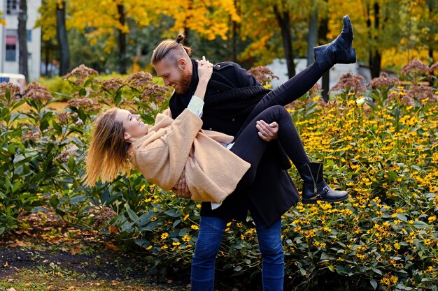 Un homme barbu rousse tient dans ses bras une jolie femme blonde dans un parc en automne.
