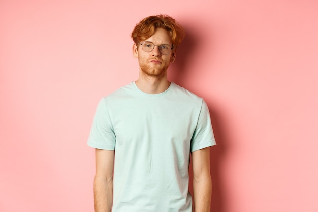 Homme barbu rousse triste et sombre en t-shirt et lunettes, regardant la caméra ennuyé et sans amusement, debout sur fond rose.