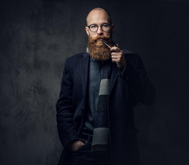 Homme barbu rousse vêtu d'un costume et de lunettes fumant une pipe traditionnelle sur fond gris foncé.