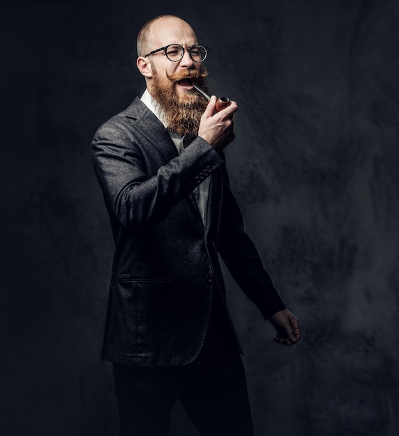 Homme barbu rousse vêtu d'un costume et de lunettes fumant une pipe traditionnelle sur fond gris foncé.