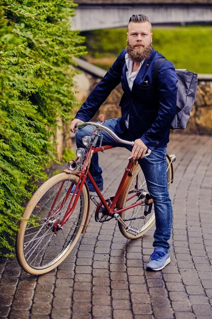 Homme barbu rousse vêtu d'une veste bleue et d'un jean sur un vélo rétro dans un parc.