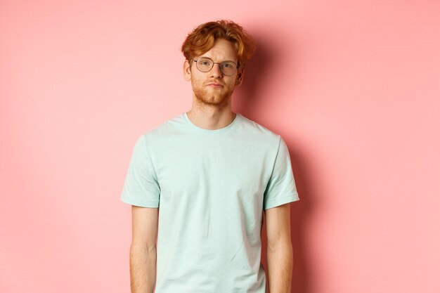 Homme barbu roux triste et sombre en t-shirt et lunettes, regardant la caméra ennuyé et sans amusement, debout sur fond rose