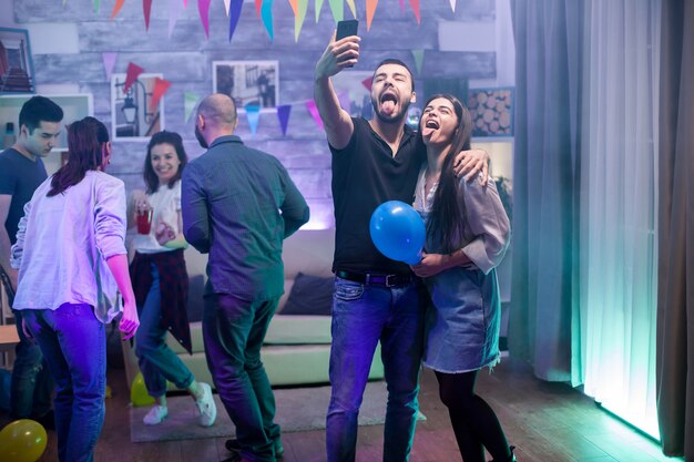 Homme barbu et sa petite amie avec la langue sortie prenant un selfie tout en faisant la fête avec leurs amis.