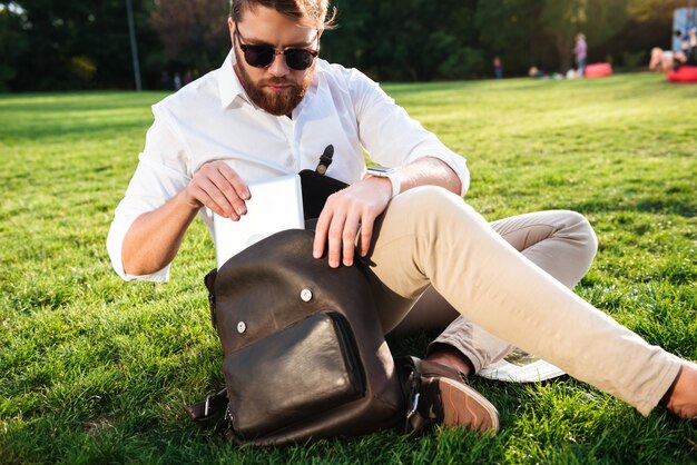 Homme barbu sérieux assis sur l'herbe à l'extérieur de l'ordinateur tablette à partir de son sac à dos
