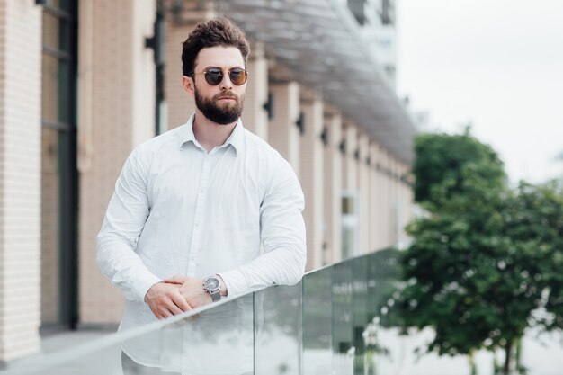 Un homme barbu, sérieux et élégant en chemise blanche et lunettes de soleil debout dans les rues de la ville près d'un bureau moderne